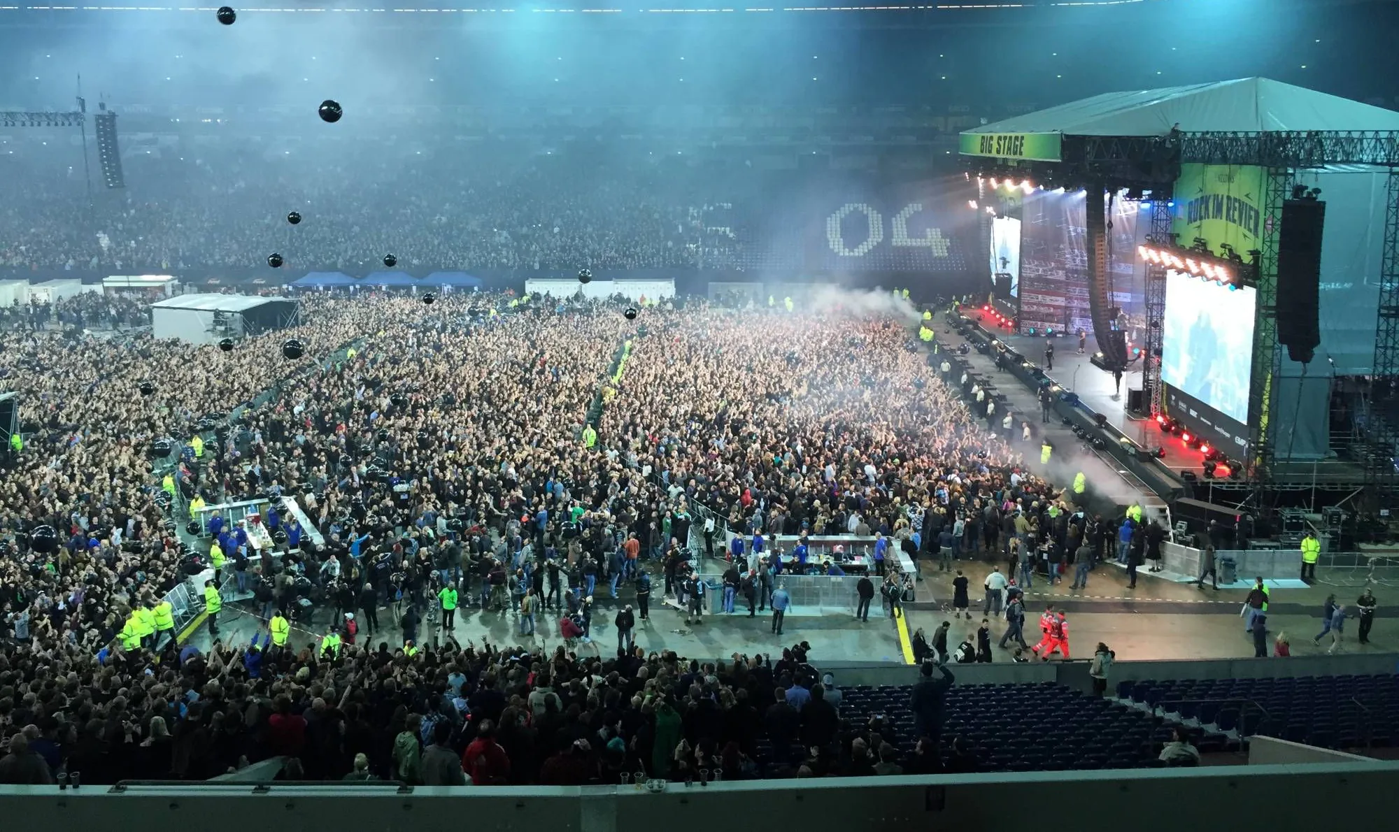 Volles Stadion beim Konzert: Publikum vor hell beleuchteter Bühne, Ordner an den Absperrungen.